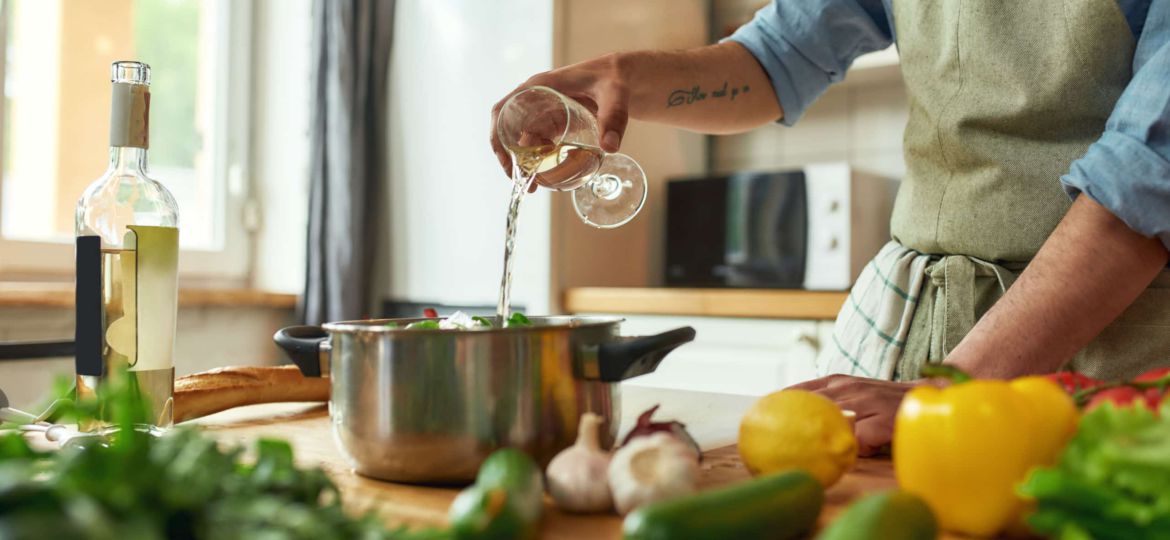 Cropped shot of man, Italian cook pouring a glass of white wine into the pan with chopped vegetables while preparing a meal in the kitchen